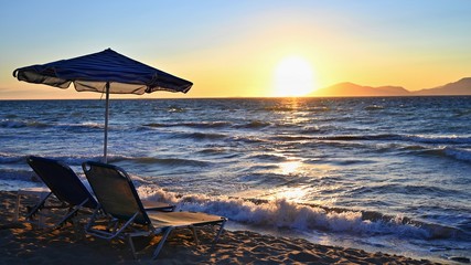 Sunbeds and umbrella on the beach at sunset by the sea. Beautiful concept for vacation, summer holidays and travel.