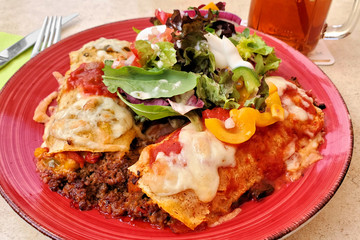 Mexican enchiladas with ground beef, peppers, cheese and salsa with a mixed salad. Close up table scene on a red plate.