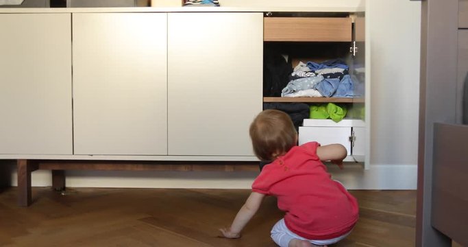 Back view of little baby opening cabinet doors and looking inside while sitting on floor in room