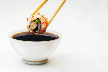Sushi held in chopsticks over a bowl with sauce on a white background