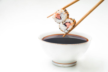 Sushi held in chopsticks over a bowl with sauce on a white background