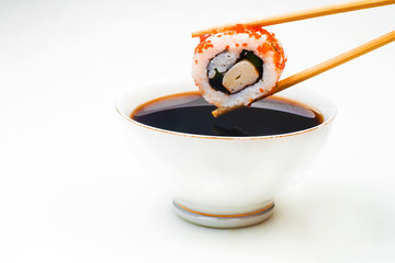 Sushi held in chopsticks over a bowl with sauce on a white background