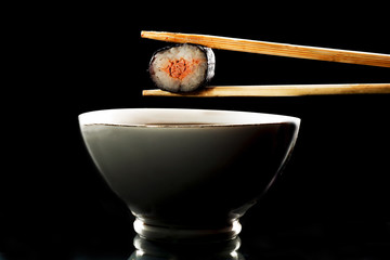 Sushi held in chopsticks over a bowl with sauce on a black background