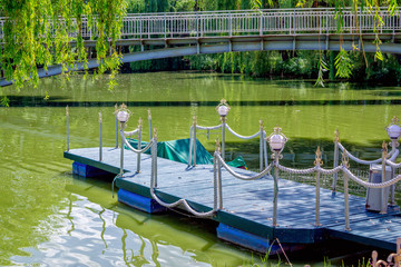 Fototapeta premium View of wooden pier, decorated with lights and rope, in calm green lake water with bridge on background