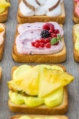 Assorted toasts with fruits and berries on wipped cream on a wood board on light background. Summer traditional homemade dessert.