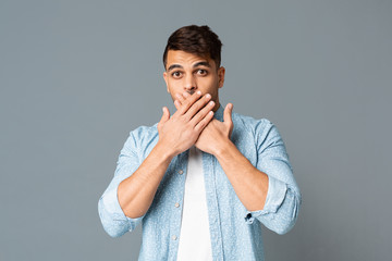 Shocked guy covering his mouth with hands on gray background.