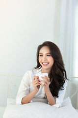 A beautiful asian woman in a white shirt and coffee cup that is smiling happily in the white bed.
