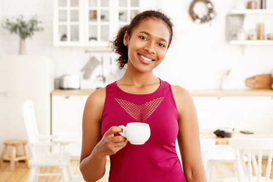 Portrait of cute African American girl with gathered hair posing in kitchen interior with cup of tea. Attractive happy dark skinned female drinking coffee, looking at camera with toothy smile