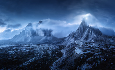 Mountains in fog at beautiful night. Dreamy landscape with mountain peaks, stones, grass, blue sky with blurred low clouds, stars and moon. Rocks at dusk. Tre Cime in Dolomites, Italy. Italian alps