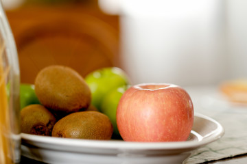 Apple, kiwi and monkey apple are put on white plate over the table in the kitchen with chair as background.