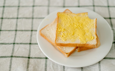 Buttered bread are put on white plate over table clothes and someone bite small piece of the bread in the corner.
