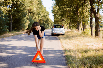 Sad student girl putting red emergency triangle stop sign because her car has broken in the countryside
