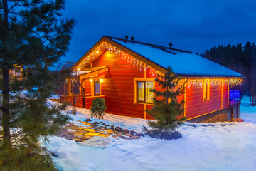 Christmas Eve. The house is decorated with garlands. Christmas holidays. Wooden house in the illumination. Winter evening. The cottage is covered with snow. Buy a house in Canada.