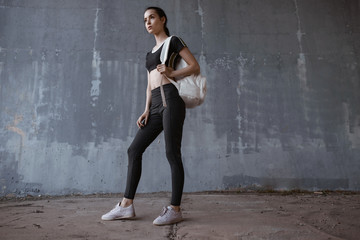 Obraz na płótnie Canvas Sports woman posing in fashion sportswear on urban gray background. Beautiful slim girl in trendy black leggings, top and white backpack.