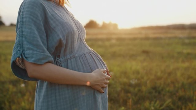 Pregnant woman dressed in linen dress and straw hat standing in meadow during sunset. Young mother to be feels happy, smiles into camera in sunlight flares, stroking and touching pregnant belly