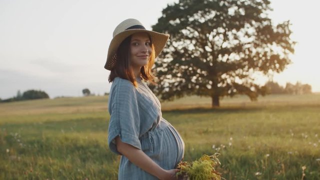 Pregnant woman walking in meadow during sunset. Mother to be dressed in linen dress and straw hat with flower bouquet in the field by oak tree takes pleasure and turns to camera in sunlight flares