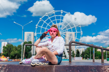 Portrait of an emotional girl in a pink cap visor wearing protective gloves and rollerblades sitting on the background of ferris wheel.