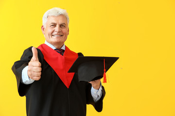 Mature man in bachelor robe showing thumb-up gesture on color background