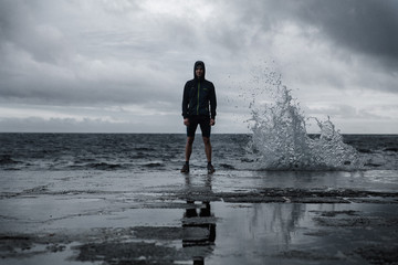 Portrait of fit man on the pier in shorts and waterproof jacket with a hood. Dramatic background with beating wave and reflection on the floor. Feeling of freedom. Sao Miguel island, Azores.