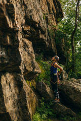 Young beautiful girl wearing in climbing equipment standing in front of a stone rock and preparing to climb against brown cracked rock and trees