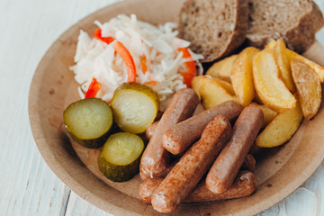 Tasty sausages with fried potatoes, cucumber, cabbage and chlobe in a paper plate on a wooden table.