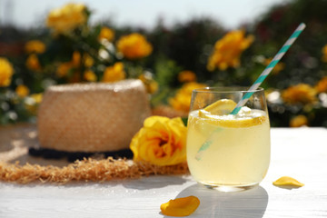 Composition with glass of refreshing lemonade and beautiful rose on white wooden table in blooming garden. Space for text