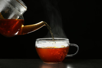 Pouring delicious hot tea into glass cup on table against dark background