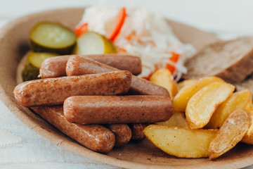 Tasty sausages with fried potatoes, cucumber, cabbage and chlobe in a paper plate on a wooden table.