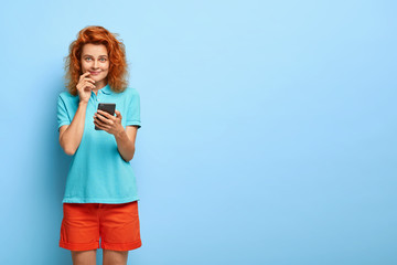 Obraz premium Satisfied red haired female blogger checks comments from followers, creats new publication, holds mobile phone, waits for email or call, wears casual outfit, stands over blue wall with blank space