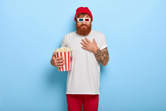 Positive carefree young man with ginger beard, touches chest, watches favourite humorous show or comedy in cinema, wears 3d glasses, holds big bucket of popcorn, dressed in fashionable outfit