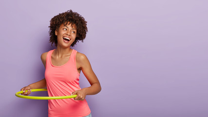 Sportive carefree woman with curly hairstyle, spins yellow plastic hoop, has gymnastic, works on figure, laughs and gazes aside, enjoys fitness activity, wears sport clothes, poses over purple wall © WHstudio Leushin N