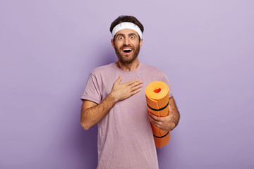 Cheerful excited sportsman looks with surprisement, touches chest, practices yoga every day, being in good physical shape, dressed in sportwear, holds orange fitness mat, isolated on purple wall