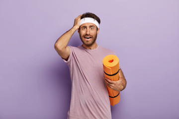 Puzzled fitness trainer touches head, holds rolled up karemat, trains in gym, shocked with little...