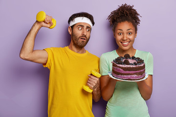 Sporty healthy lifestyle against junk food. Puzzled sportsman holds dumbbells, has fitness...