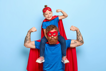 Strong powerful dad and little female child on his shoulders show muscles, ready to defend you and struggle, dressed in superman clothes, isolated on blue background, prentend having superhuman power