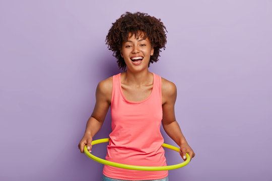 Photo of optimistic dark skinned woman rotates hule hoop, aims to have perfect waist, smiles broadly, likes sport and gymnastics, has fun during exercising, wears casual outfit. Loosing weight concept