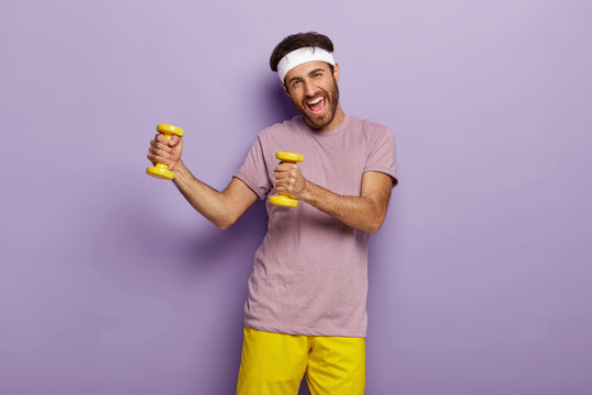 Hardy positive man has training with two yellow weights, excellent physical shape, glad expression, wears casual outfit, isolated on purple background, demonstrates how to do exercise correct
