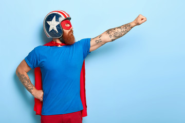 Profile shot of serious bearded man makes flying gesture, looks into distance, wears protective helmet and red cape, poses against blue background with empty space for your promotional content