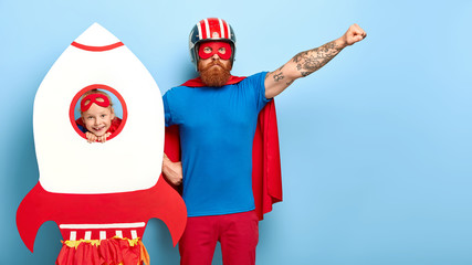 Serious bearded father raises arm, pretends being superhero, wears helmet, mask, cape and blue t...