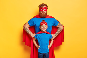 Photo of confident ginger father and daughter keep hands on waists, pretend being superhero and defend people, wear special costumes, isolated on yellow background. Family team ready to work © WHstudio Leushin N