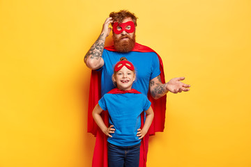 Playful father with small daughter wear superman costumes, play together at home, confused dad scratches head with bewilderment, enjoy togetherness, isolated on yellow wall. Chilhood, parenthood