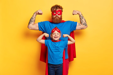 Superhero family. Strong father and small child show biceps, pretend being superman, wear red masks, blue t shirts and cloaks, motivated for successful future, stand agaisnt yellow studio wall - obrazy, fototapety, plakaty