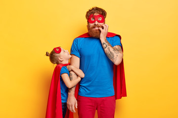 Nervous worried bearded father organizes real holiday for children, little girl embraces his hand, wear superhero suits, stand against yellow studio background. Parenthood, fatherhood concept. © WHstudio Leushin N