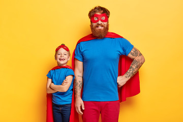 Studio shot of cheerful family in superhero suits, look confidently at camera, express their...