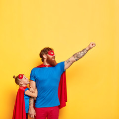 Superhero man spends leisure time with small child, makes flying gesture, raises fist, wear...