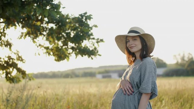 Pregnant woman dressed in linen dress and hat standing by oak tree in meadow outdoors during sunset. Beautiful mother to be relax stroking pregnant belly smiles into camera in evening sunlight