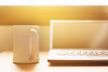 laptop is on the desk at home near window in the morning. there is a cup of coffee for refresh you from awake. the sun is shining from outside onto the desk.
