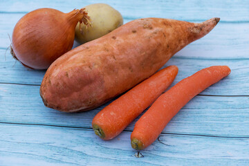 Ripe carrots, potato, sweet potato and brown onion on a blue wooden table background. Cooking ingredients. Harvest