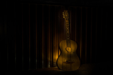 An wooden acoustic guitar is against a grunge textured wall. The room is dark with a spotlight for...
