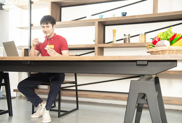 Asian smiling young man with casual  red t-shirt enjoy having breakfast, eating sandwich, Young man cooking food and drink in the loft style kitchen room